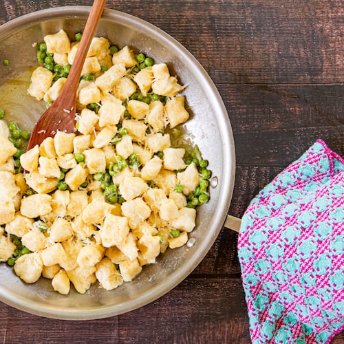 A large metal mixing bowl holds a batch of ricotta gnocchi and peas, sprinkled with Parmesan cheese. A wooden spoon is tucked into the bowl, and a cloth napkin is artfully arranged next to it.