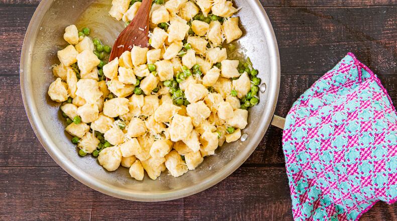 A large metal mixing bowl holds a batch of ricotta gnocchi and peas, sprinkled with Parmesan cheese. A wooden spoon is tucked into the bowl, and a cloth napkin is artfully arranged next to it.