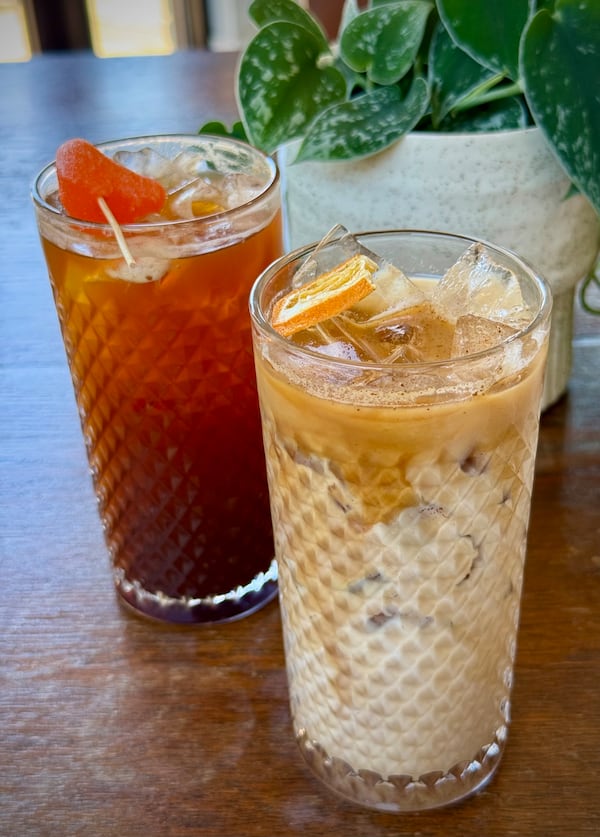 Two tall glasses filled with iced coffee, the glass on the left garnished with a piece of candy that looks like an orange segment and the glass on the right with a milkier color and a dehydrated orange slice garnish.
