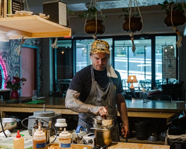 Chef Chris McCord stands at a wooden work table stirring something in a tall metal bowl. There are sauces in plastic squeeze bottles in the foreground and other ingredients on the counter.