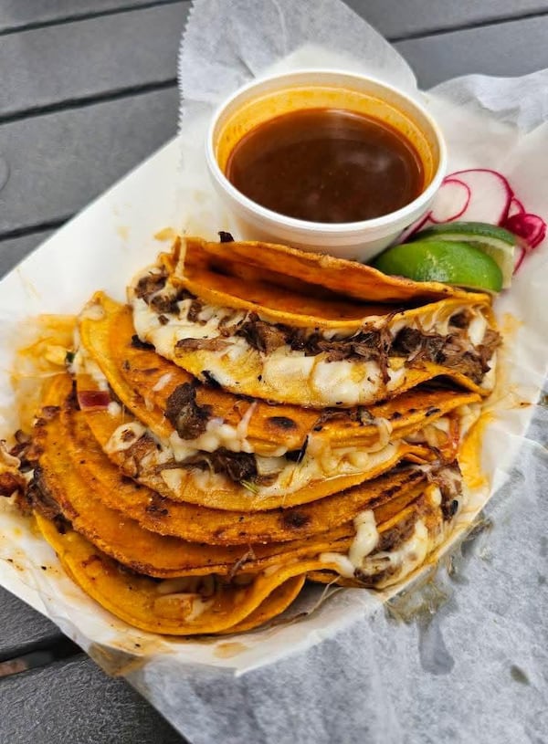 A paper boat dish filled with a pile of birria tacos, a dipping sauce and lime wedges.