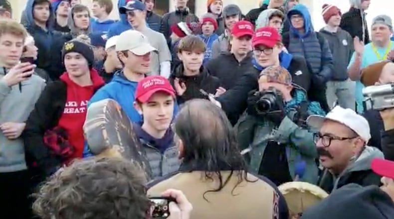 In this Friday, Jan. 18, 2019 image made from video provided by the Survival Media Agency, a teenager wearing a "Make America Great Again" hat, center left, stands in front of an elderly Native American singing and playing a drum in Washington.
