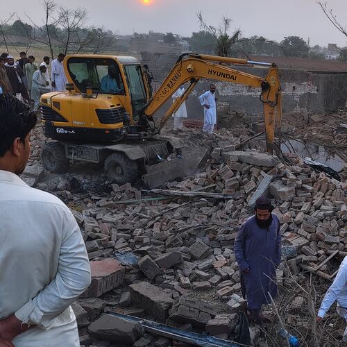 A worker clear rubble with an excavator as a police officer and local residents gather at the site of overnight suicide bombing at a security post in Fatah Khel, in Bannu, a district in Pakistan's Khyber Pakhtunkhwa province bordering Afghanistan, Sunday, May 10, 2026. (AP Photo/Aamad Khattak)