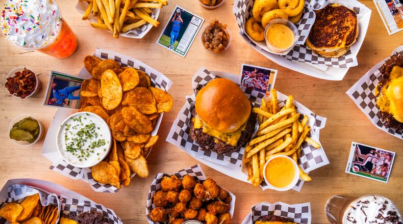 An overhead shot of a table full of food from a burger restaurant, including burgers, fries, frozen sodas, tater tots, chips and dip and sides and other accouterments.
