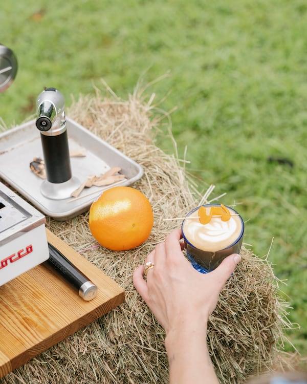 A hand reaches for a cortado with a lemon peel speared across it. The drink sits atop a bale of hay with an orange next to it.