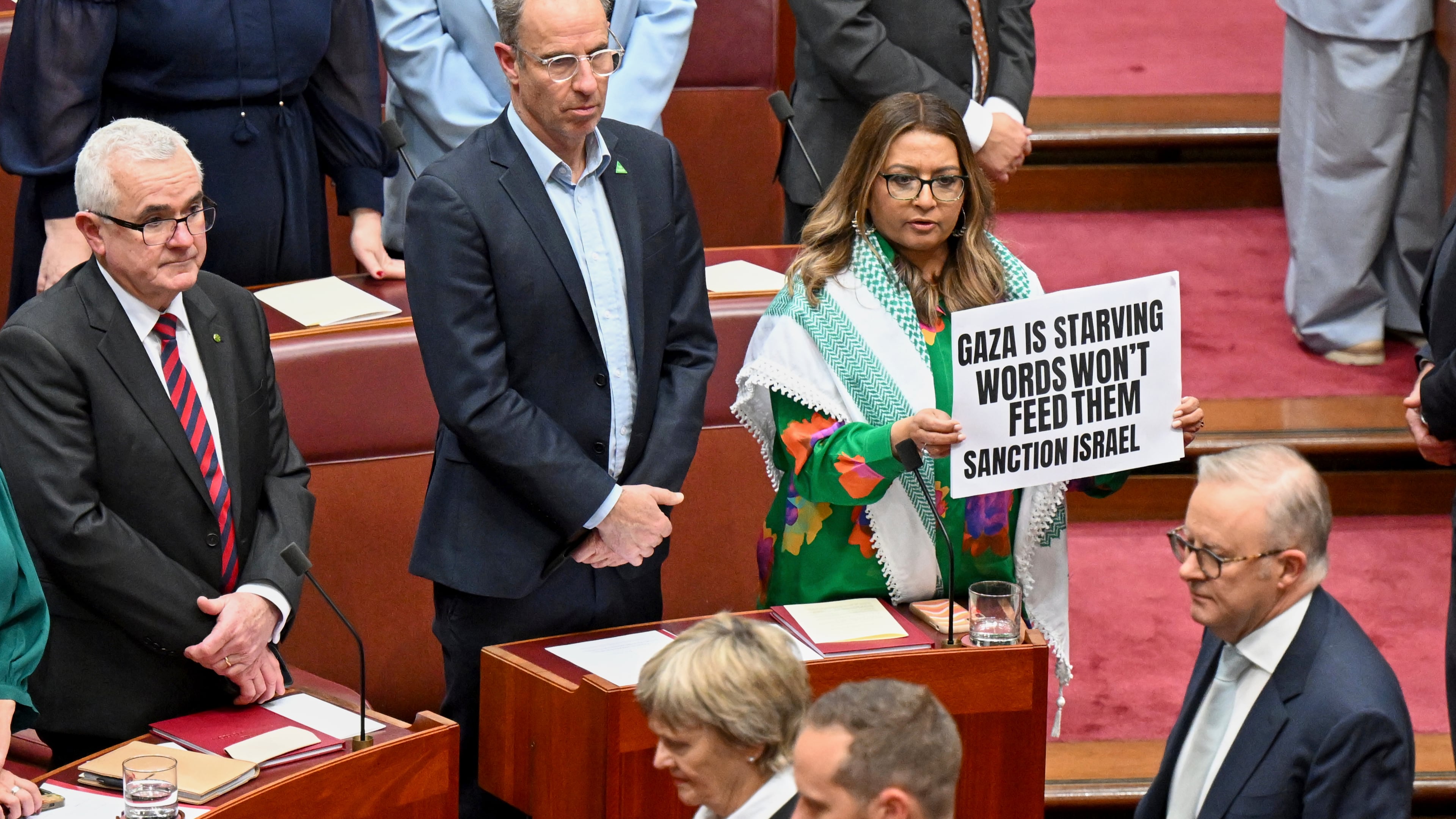Greens Senator Mehreen Faruqi holds a placard as Australian Prime Minister Anthony Albanese walks in the Senate chamber for the opening of the 48th Federal Parliament at Parliament House in Canberra, Australia, Tuesday, July 22, 2025. (Lukas Coch/AAP Image via AP)