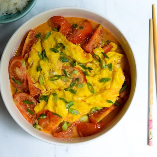 A serving bowl holds tomatoes and eggs garnished with chopped scallions. Next to the bowl are two smaller bowls of steamed white rice and a pair of chopsticks.