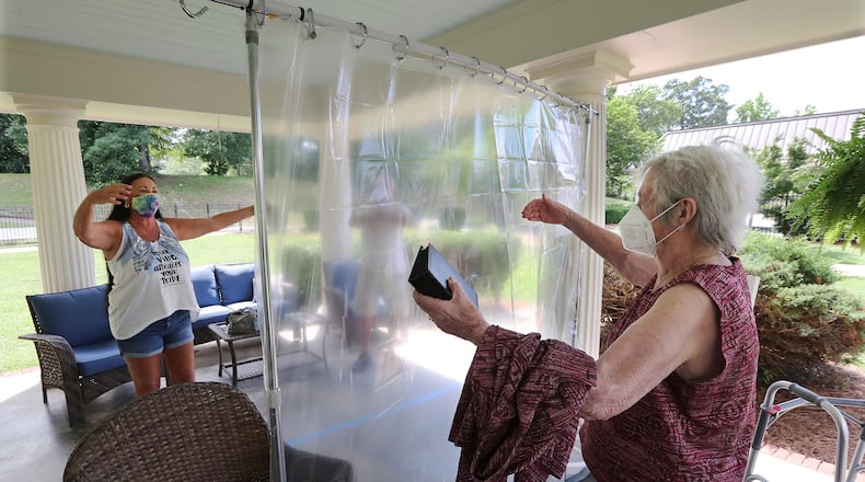 Elsie Grant, 86, sets her walker aside reaching for an air hug from her daughter Wanda Schroeder, socially distanced behind a protective plastic curtain when families could visit loved ones in the outdoor pavilion at Westbury Medical Care & Rehab earlier this month. CURTIS COMPTON / CCOMPTON@AJC.COM