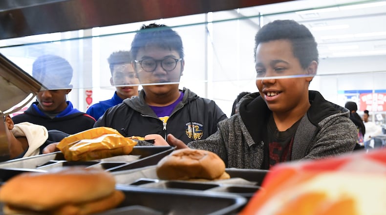 Eighth grader Elijah Caraballo, 14, reaches for a tray next to the hamburgers and Cheetos, as Alexander Boualavong (left), 15, waits his turn during lunch in the cafeteria at Elite Scholars Academy in Jonesboro. The school is in Clayton County, where all students get a free lunch so there is no meal debt. JOHN AMIS / FOR THE AJC