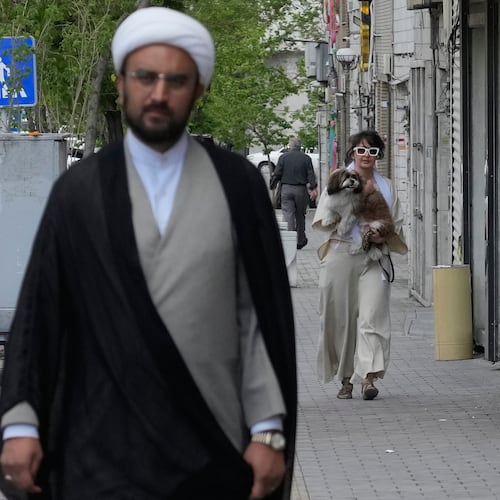 A woman carries her dog as a cleric walks on a sidewalk in Tehran, Iran, Tuesday, April 21, 2026. (AP Photo/Vahid Salemi)