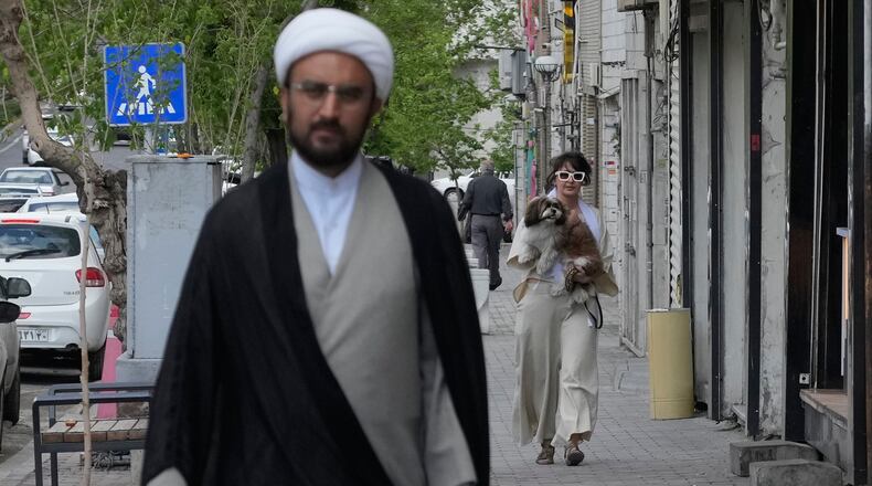 A woman carries her dog as a cleric walks on a sidewalk in Tehran, Iran, Tuesday, April 21, 2026. (AP Photo/Vahid Salemi)
