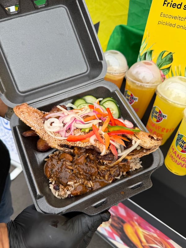 A styrofoam take out container contains a fried, whole red snapper and rice with oxtail gravy. There are also four plastic cups filled with drinks in the image.