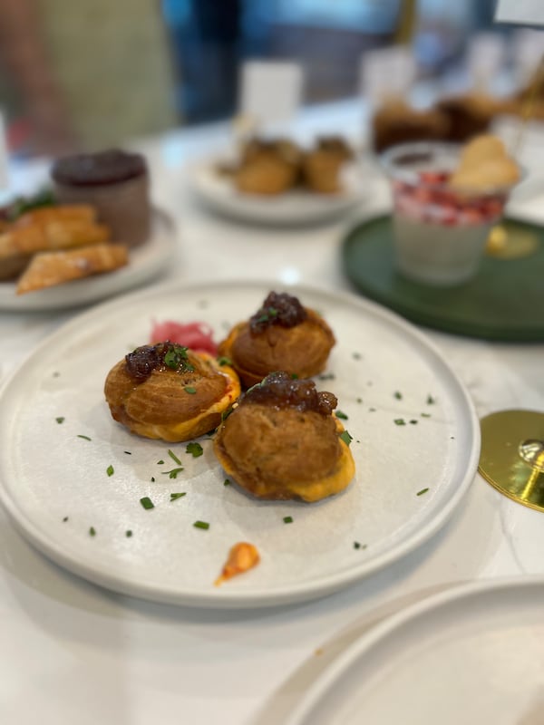 Three profiteroles filled with pimento cheese and garnished with bacon jam are on a plate. The plate is on a white countertop that looks like it could be in the Galette's storefront because there are plates of other bakery items also on display.