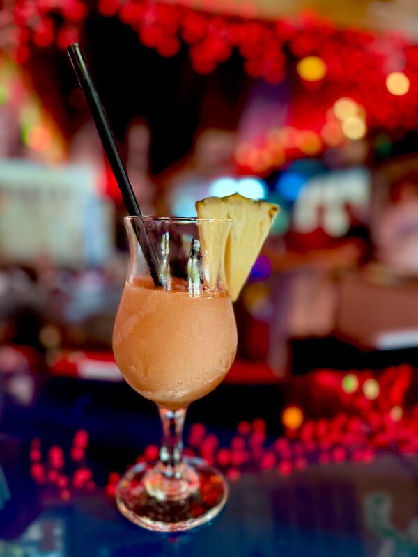 A hurricane frozen cocktail is garnished with a large pineapple wedge and a cocktail stirrer. It's placed on a bar, with festive out-of-focus lights in the background.