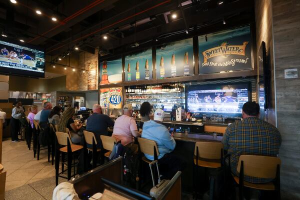 All of the bar chairs are filled (except for one) with patrons in this photo of Atlanta Bread and Bar. There is a tv showing a sporting event and rows of liquor bottles on the wall behind the bar.