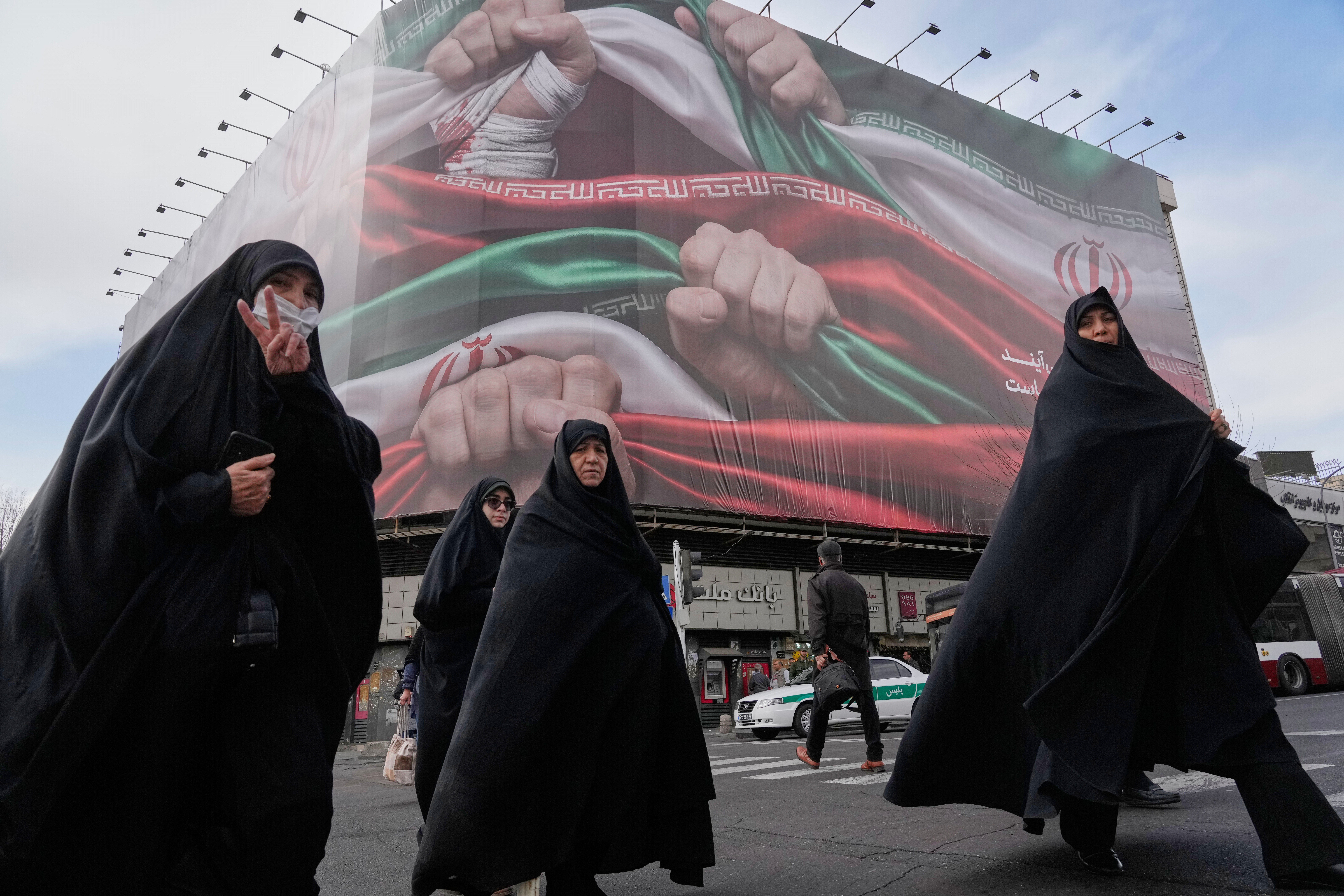FILE - Women, one flashing a victory hand gesture, cross a street under a huge banner showing hands firmly holding Iranian national flags as a sign of patriotism, in Tehran, Iran, Jan. 14, 2026. (AP Photo/Vahid Salemi, File)