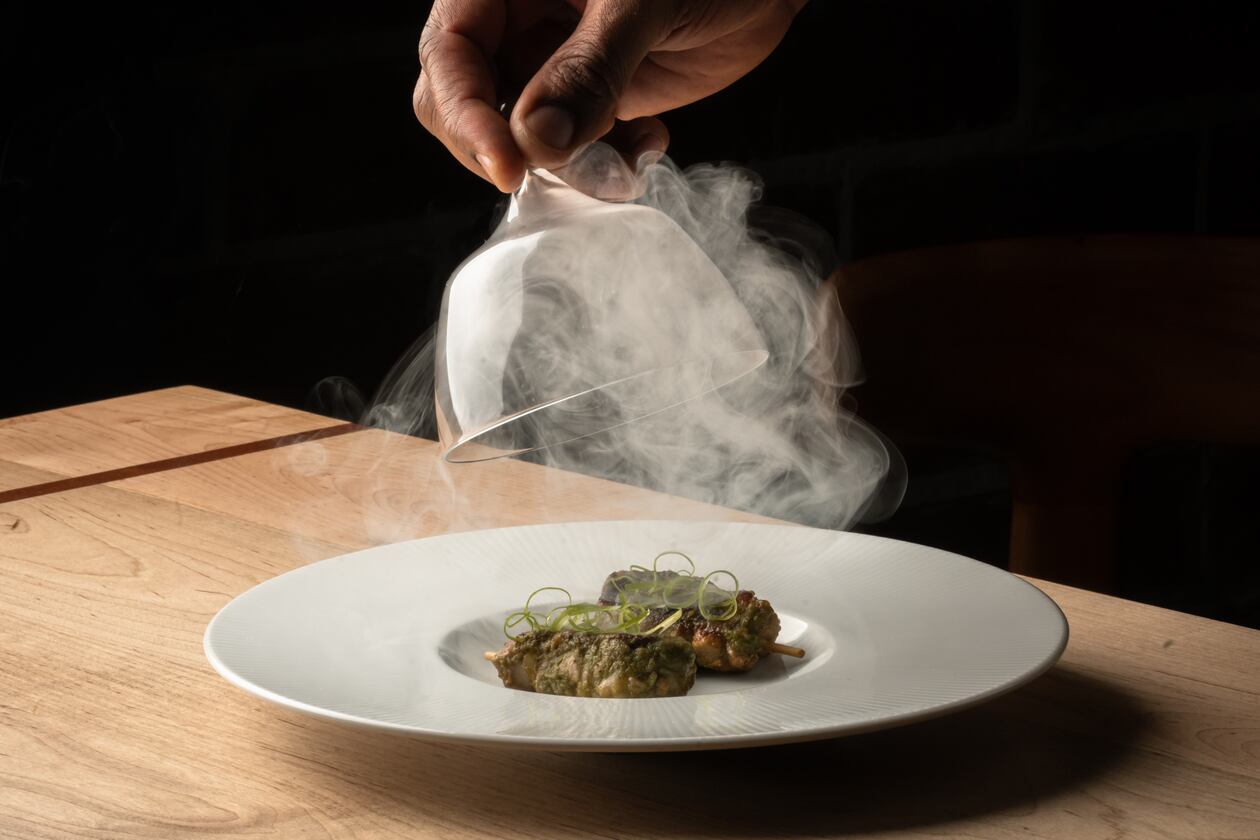 A hand lifts a glass cloche off a plate of food, releasing smoke into the air in a darkened room.