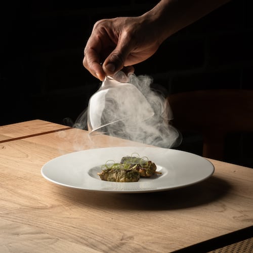 A hand lifts a glass cloche off a plate of food, releasing smoke into the air in a darkened room.