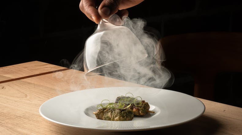 A hand lifts a glass cloche off a plate of food, releasing smoke into the air in a darkened room.