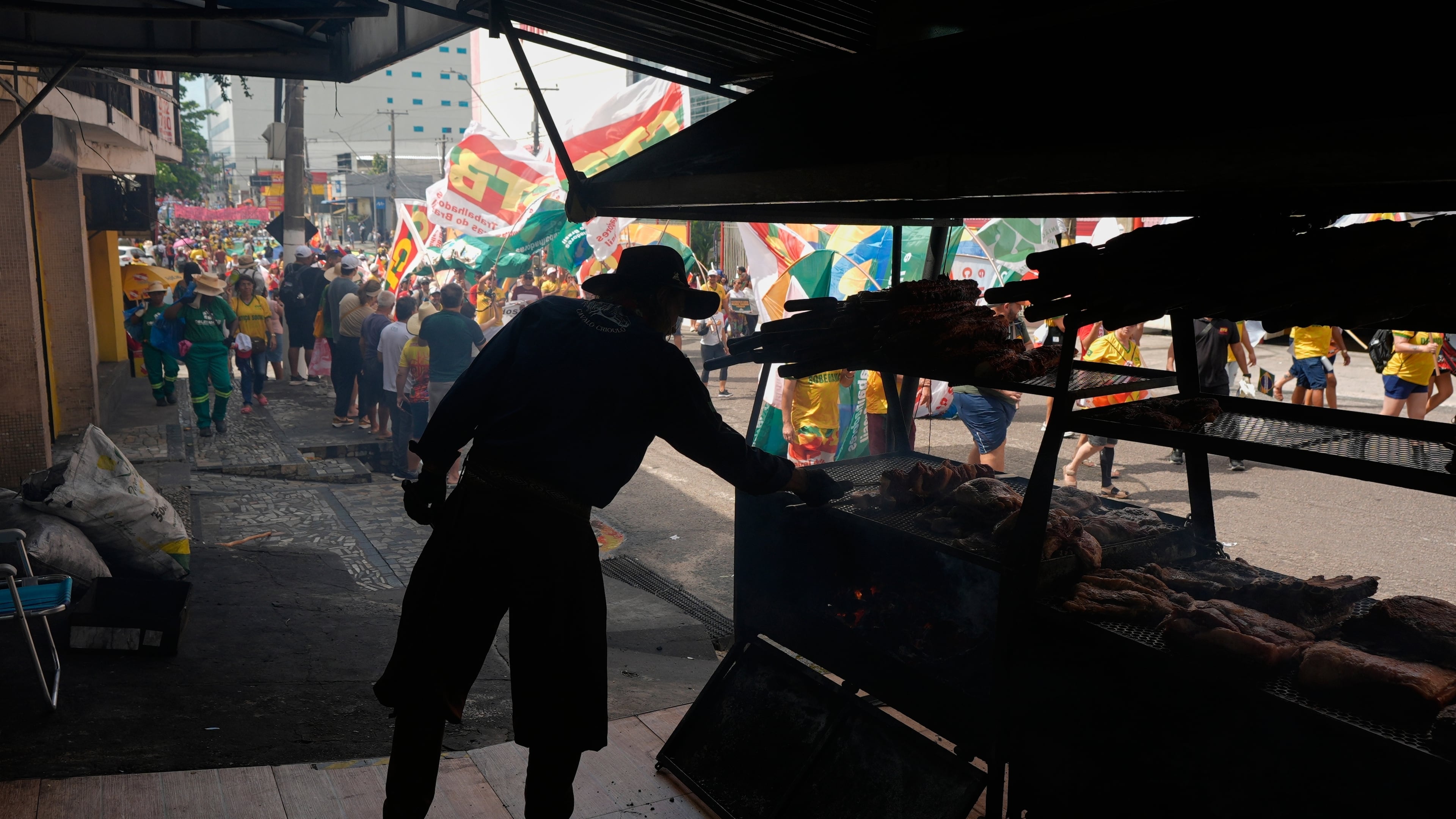 A vender cooks meat as a climate protest goes by during the COP30 U.N. Climate Summit, Saturday, Nov. 15, 2025, in Belem, Brazil. (AP Photo/Fernando Llano)