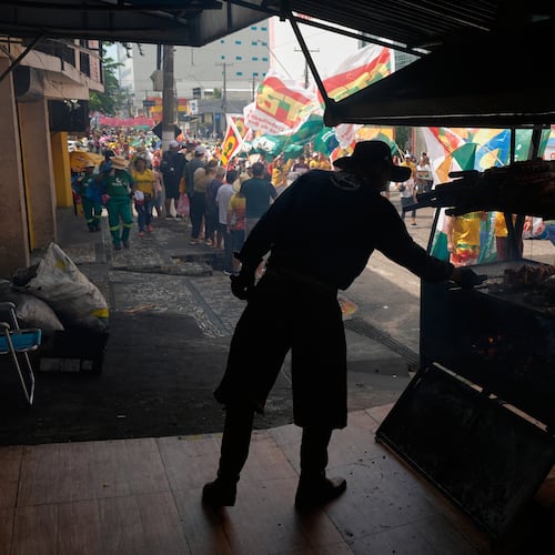 A vender cooks meat as a climate protest goes by during the COP30 U.N. Climate Summit, Saturday, Nov. 15, 2025, in Belem, Brazil. (AP Photo/Fernando Llano)