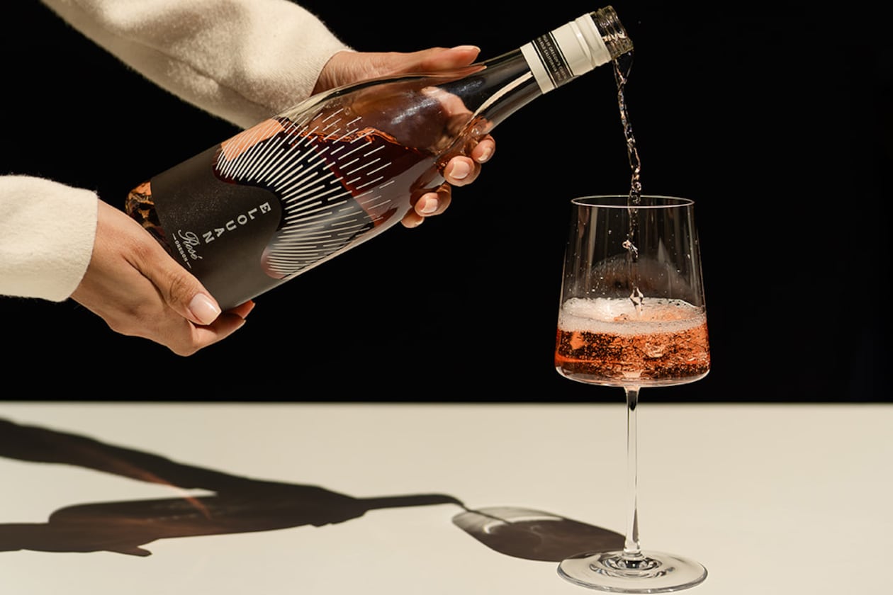 Two hands are shown pouring a glass of Elouan Rose wine in a stylized photo. The person pouring the wine is not seen, but her arms are clothed in an elegant long-sleeve shirt, and her nails are tastefully manicured, suggesting she is a server at a fancy restaurant.