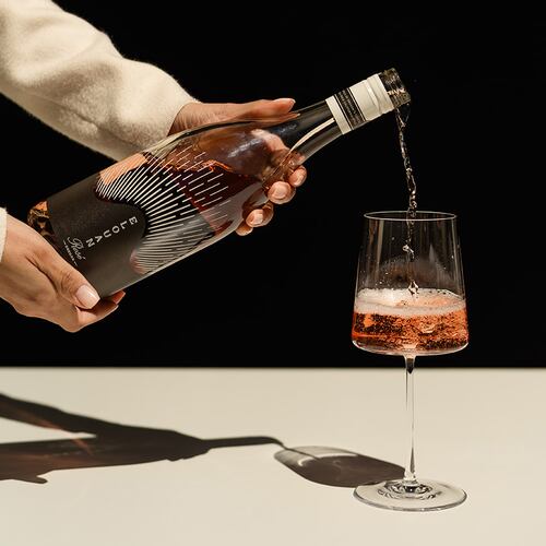 Two hands are shown pouring a glass of Elouan Rose wine in a stylized photo. The person pouring the wine is not seen, but her arms are clothed in an elegant long-sleeve shirt, and her nails are tastefully manicured, suggesting she is a server at a fancy restaurant.