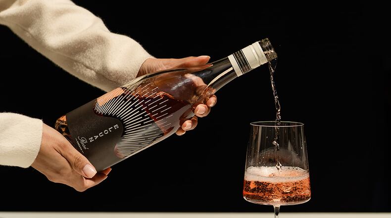 Two hands are shown pouring a glass of Elouan Rose wine in a stylized photo. The person pouring the wine is not seen, but her arms are clothed in an elegant long-sleeve shirt, and her nails are tastefully manicured, suggesting she is a server at a fancy restaurant.