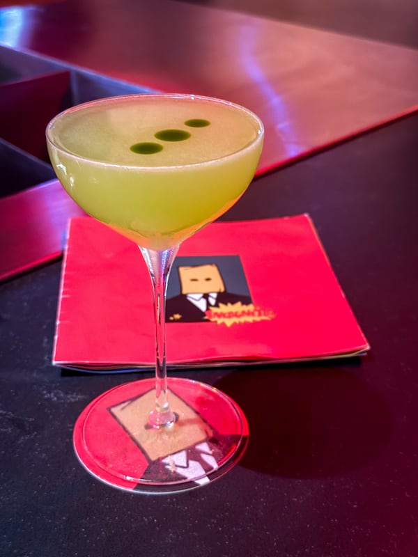 A cocktail in a coupe glass sits on a black bar with three drops of green oil on its surface.