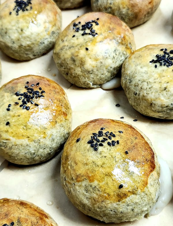 Rows of matcha buns are symetrically arranged. The buns appear to be garnished with small seeds, and some have a little frosting or other creamy substance oozing out from the bottom.