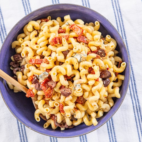 A plastic serving bowl of corkscrew-shaped pasta made with sliced sun-dried tomatoes, blue cheese and Kalmata olives is shown on top of a kitchen towel. The stripe on the towel is the same color as the serving bowl. A wooden spoon is tucked into the bowl.