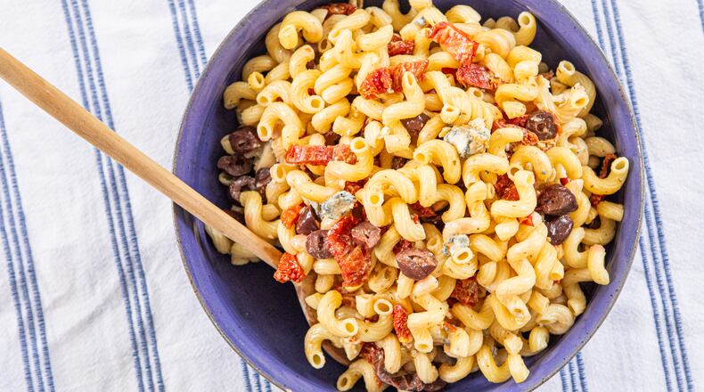 A plastic serving bowl of corkscrew-shaped pasta made with sliced sun-dried tomatoes, blue cheese and Kalmata olives is shown on top of a kitchen towel. The stripe on the towel is the same color as the serving bowl. A wooden spoon is tucked into the bowl.
