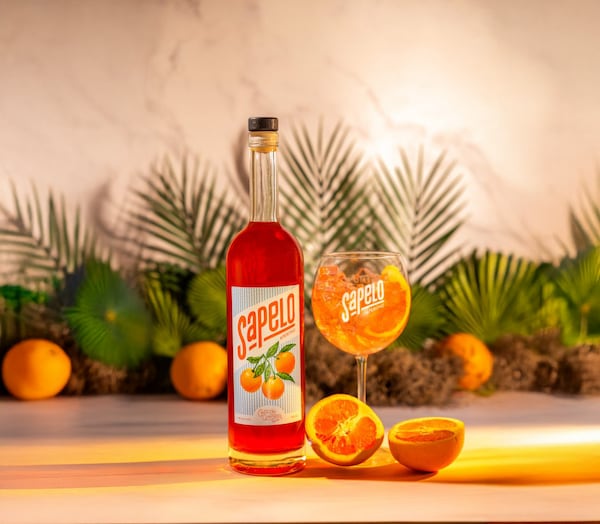 A bottle of orange liqueur next to large, round wine glass holding a spritz cocktail, with a halved orange in front of the glass and palm fronds and more oranges in the background.