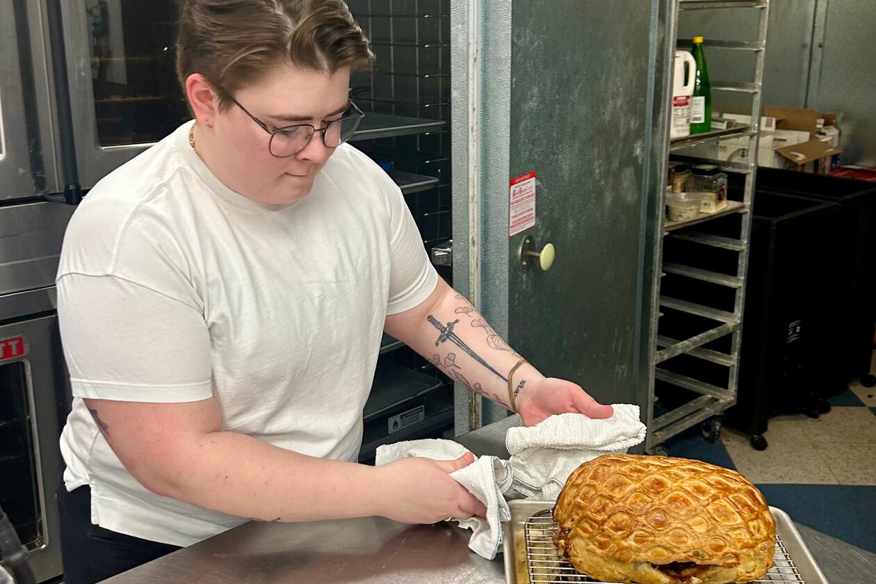 Hannah Chandler wearing a white t-shirt and glasses, holding a freshly cooked porchetta roast on a wire rack in a restaurant kitchen.