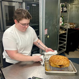 Hannah Chandler wearing a white t-shirt and glasses, holding a freshly cooked porchetta roast on a wire rack in a restaurant kitchen.