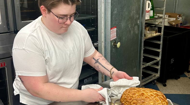 Hannah Chandler wearing a white t-shirt and glasses, holding a freshly cooked porchetta roast on a wire rack in a restaurant kitchen.