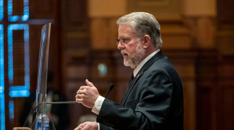 side photo of white man with grey hair and glasses, raising hand as he speaks
