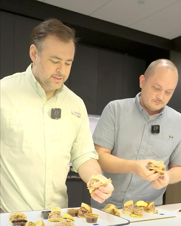 AJC Food Critic Henri Hollis and AJC Video Operations Manager John Kushnerick each pick up the first contender for Best Pulled Pork Sandwich from trays that showcase all six contenders.
