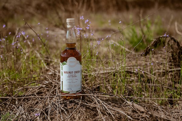 A bottle of whiskey is placed on the ground in a dry field amid twigs and sparce grass.