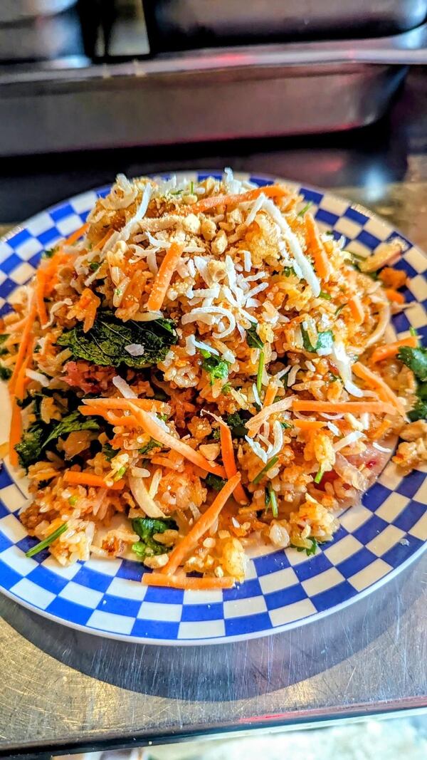 Blue and white checkered plate with crispy rice, shredded carrot, green herbs, grated coconut set on top of a metal prep table.
