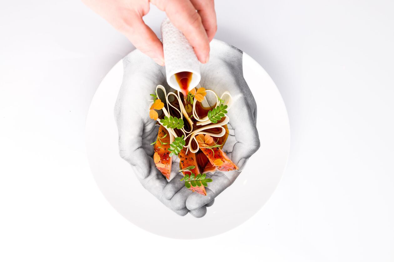 A hand pours a pitcher of sauce over a daikon and hamachi crudo salad that's nestled into a plate with an image of cupped hands on the bottom.