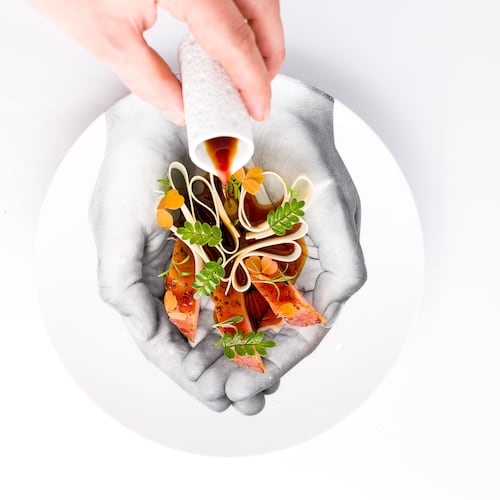 A hand pours a pitcher of sauce over a daikon and hamachi crudo salad that's nestled into a plate with an image of cupped hands on the bottom.