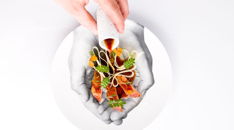 A hand pours a pitcher of sauce over a daikon and hamachi crudo salad that's nestled into a plate with an image of cupped hands on the bottom.