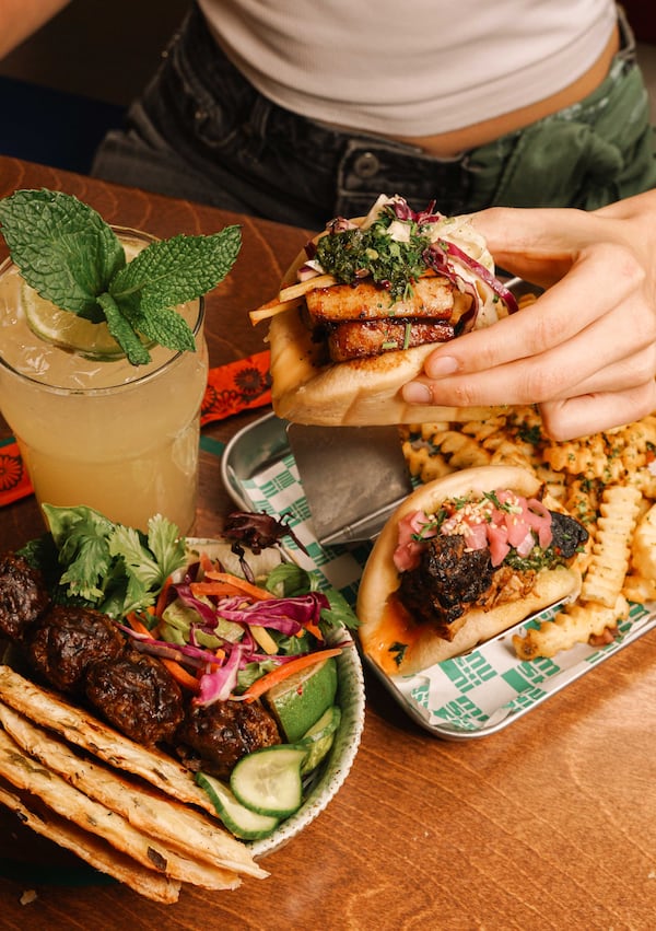 A woman's hand lifts a bao bun from the plate in front of her, which also holds another bao bun and french fries. There's also a large cocktail in a pint glass and a dish of beef meatballs with veggies and scallion pancakes.