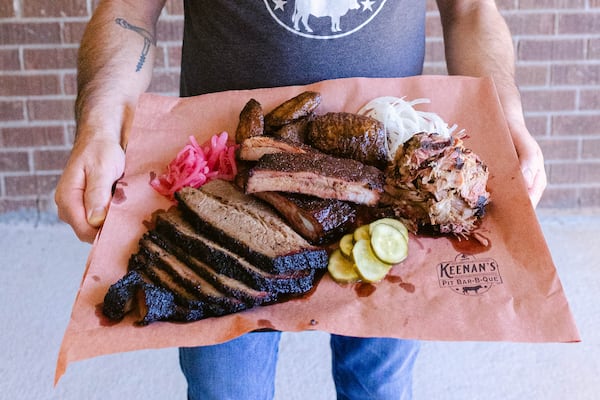 A man extends a tray towards the camera. The tray is covered in paper labeled with the Kennan's logo. It contains brisket, ribs, smoked meat and potatoes and is garnished with pickle slices and pickled red onions.