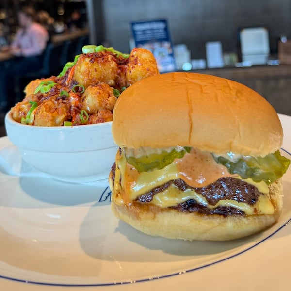 A loaded double cheeseburger from NFA burger is accompanied by a bowl of loaded tater tots from Roshambo. They are placed next to each other on a wood-topped table in a restaurant environment.