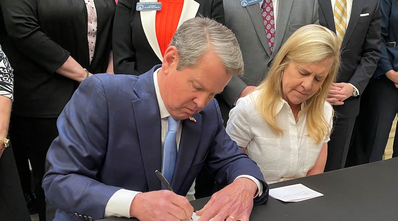 Gov. Brian Kemp signs SB 65, a bill to establish a state-based exchange under the Affordable Care Act, where Georgians could shop for ACA health insurance plans on a state-run website.  If approved by the Biden administration, people who now shop on the federal healthcare.gov website would instead shop on a state-run website. Kemp is flanked by his wife, Marty, and sits before a crowd of legislators at the Georgia Capitol on Tuesday, May 2, 2023. (PHOTO by Ariel Hart)
