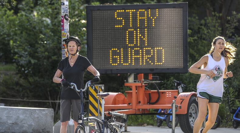 People wait for the light to change at Monroe Drive and 10th Street in Atlanta at the Beltline on Tuesday, May 5, 2020. A sign warns of the need for continuing vigilance against the coronavirus. JOHN SPINK/JSPINK@AJC.COM