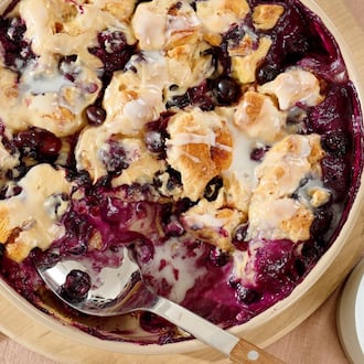 An overhead shot of a blueberry buckle in a beige casserole dish with a serving spoon in it and a cup of coffee next to it.