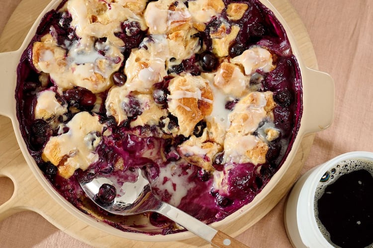 An overhead shot of a blueberry buckle in a beige casserole dish with a serving spoon in it and a cup of coffee next to it.